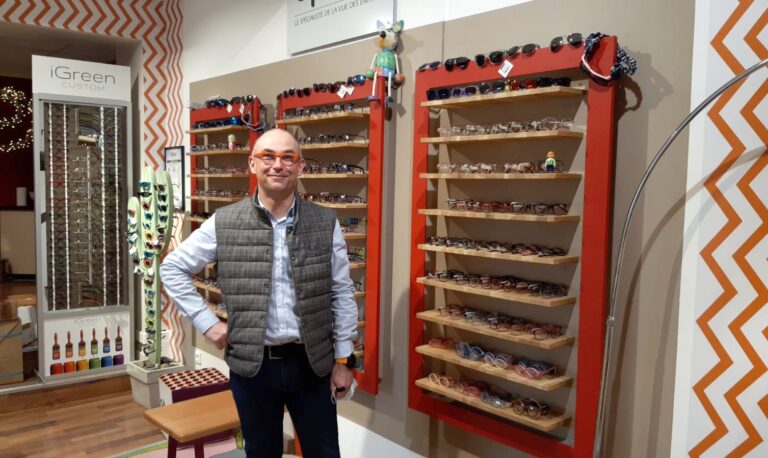 opticien qui pose devant des rayons de lunettes de vue