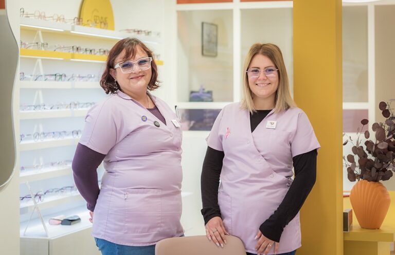 deux opticiennes souriantes à lunettes posent dans leur magasin d'optique. Elles portent toutes les deux une blouse lila identique
