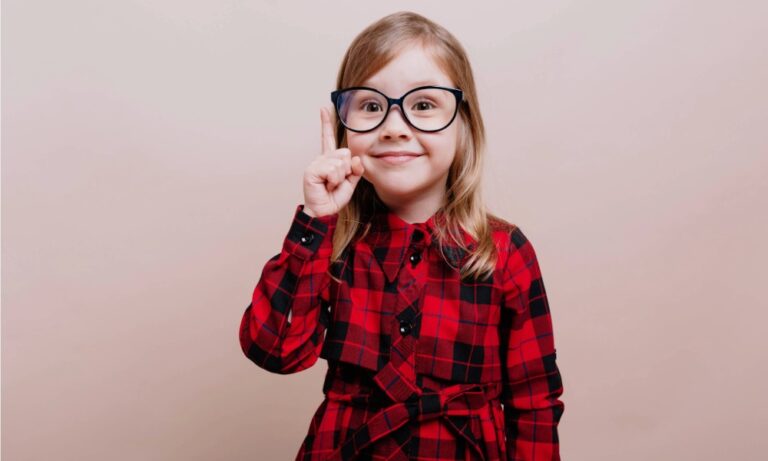 petite fille souriante avec de grandes lunettes de vue noires qui pointe son doigt vers le haut